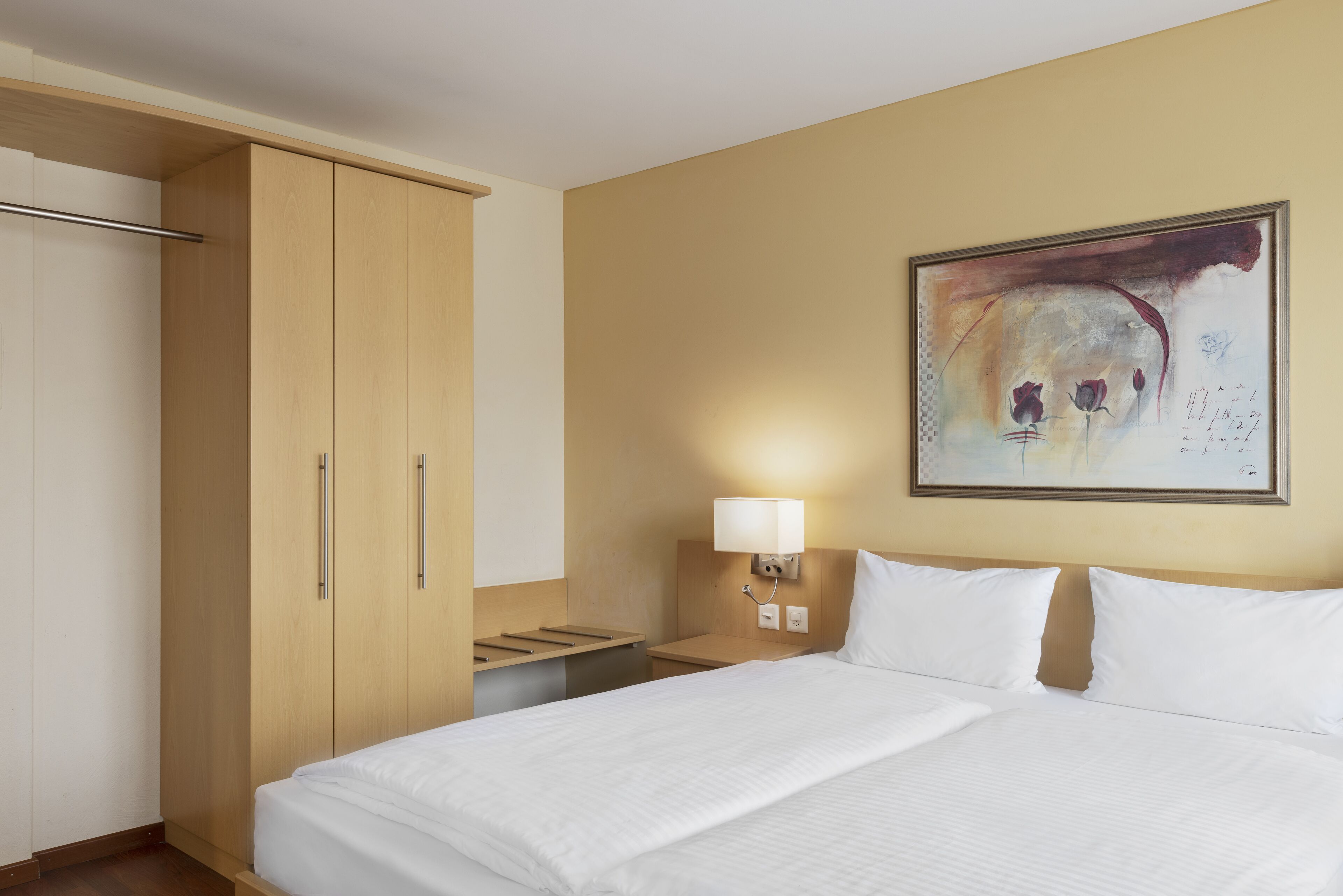 Una habitación de hotel con cama, mesita de noche, lámpara, un cuadro en la pared y armario.