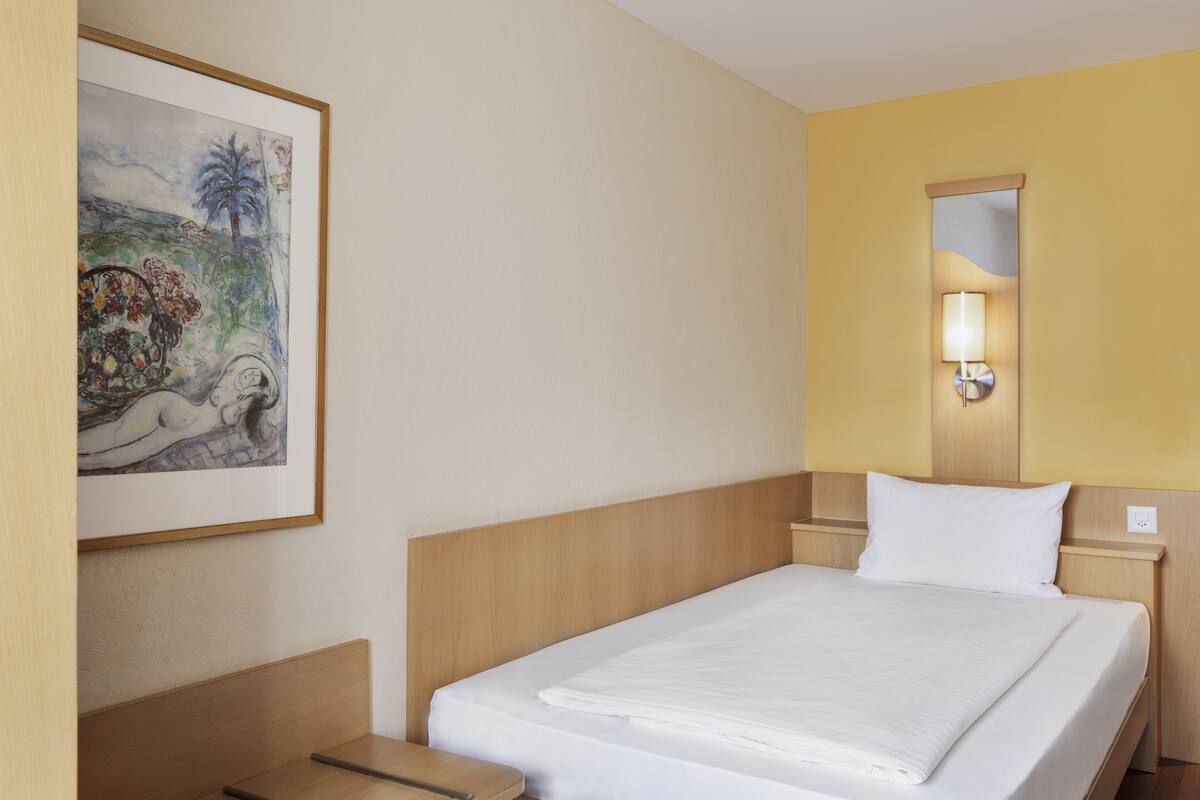 A hotel room with a neatly made bed, a bedside table, a nightstand, a wall-mounted lamp, and a framed painting on the wall.