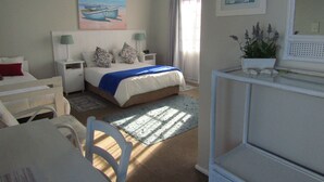 Une chambre à coucher comprenant un lit, des tables de chevet, un bureau, une chaise, une fenêtre avec des rideaux, un tableau au mur et une plante en pot.