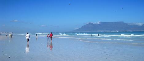 A beach with people walking and standing by the water, a clear sky, and distant mountains.