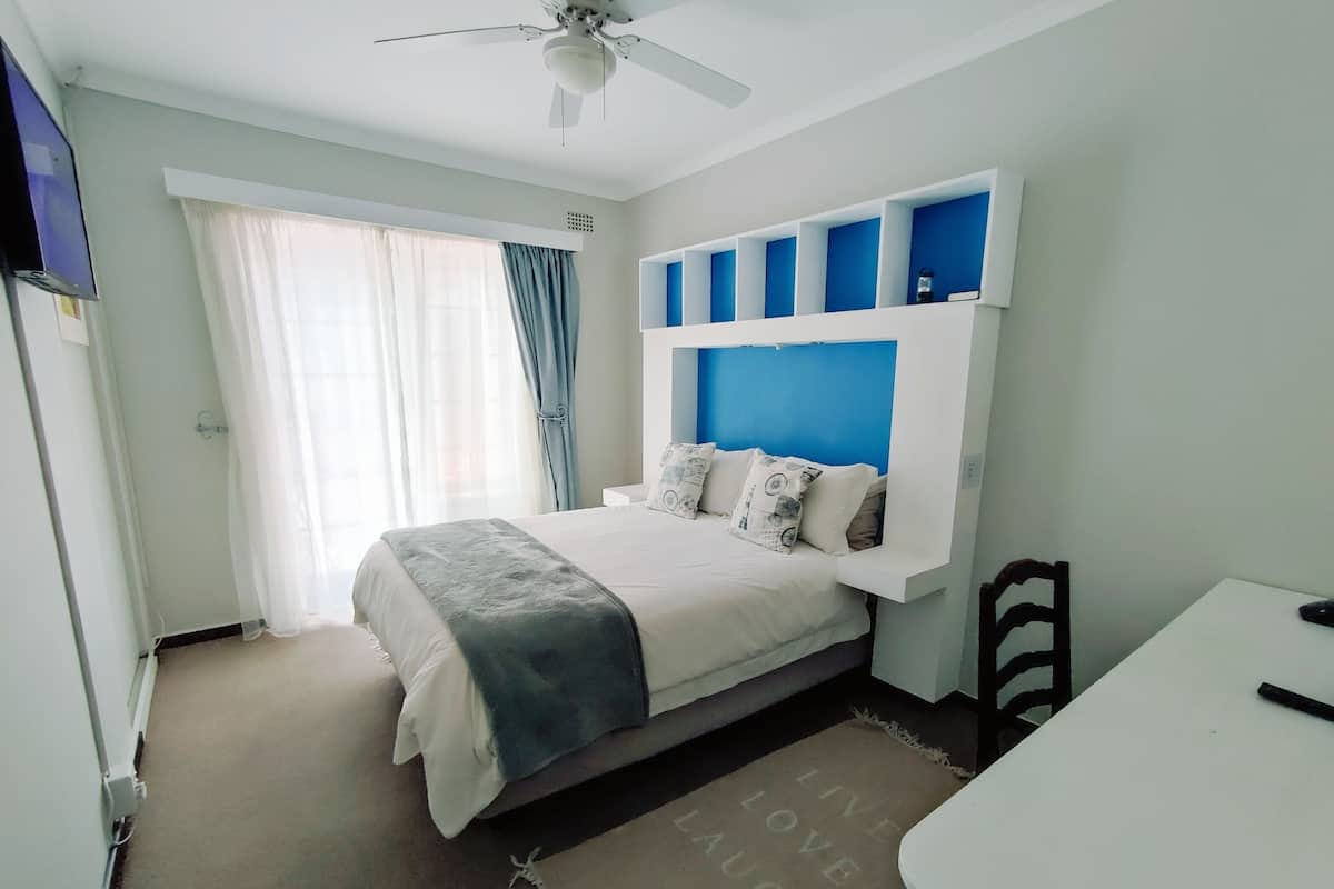 A bedroom with a bed, a desk, a chair, a ceiling fan, and a window with curtains.