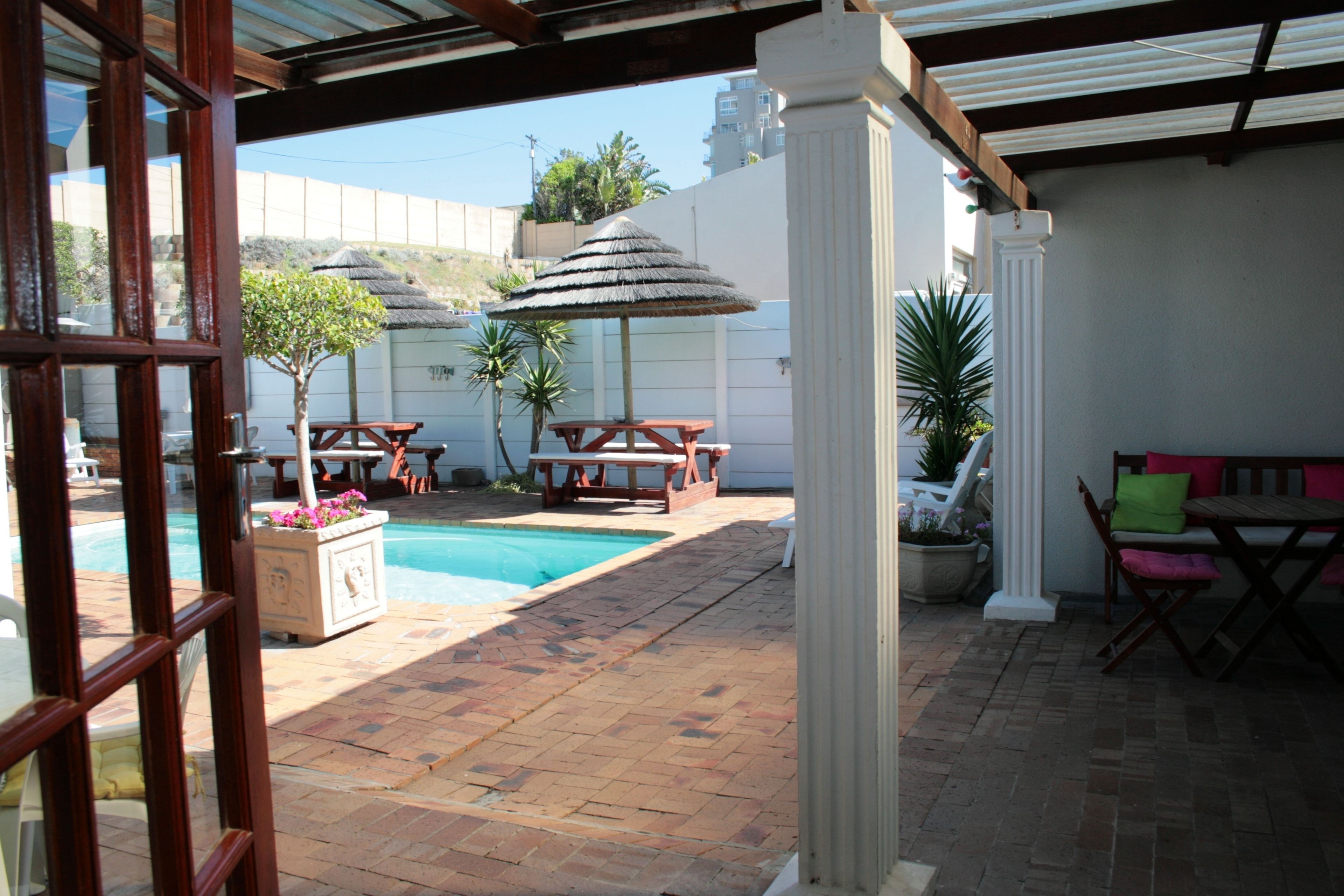 Um pátio externo com piscina, mesas e cadeiras de madeira, e plantas em vasos.