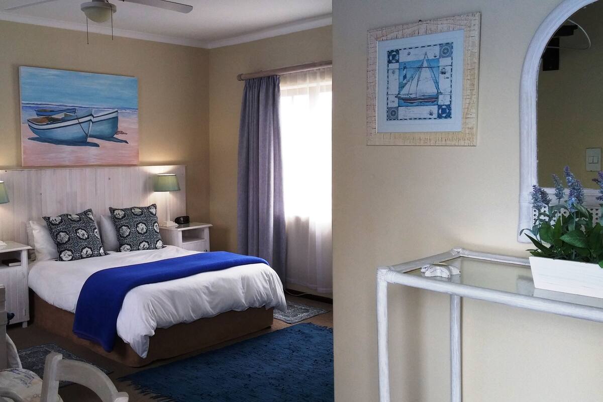 A hotel room with a bed, two bedside tables with lamps, a painting of a boat, a window with curtains, and a mirror.