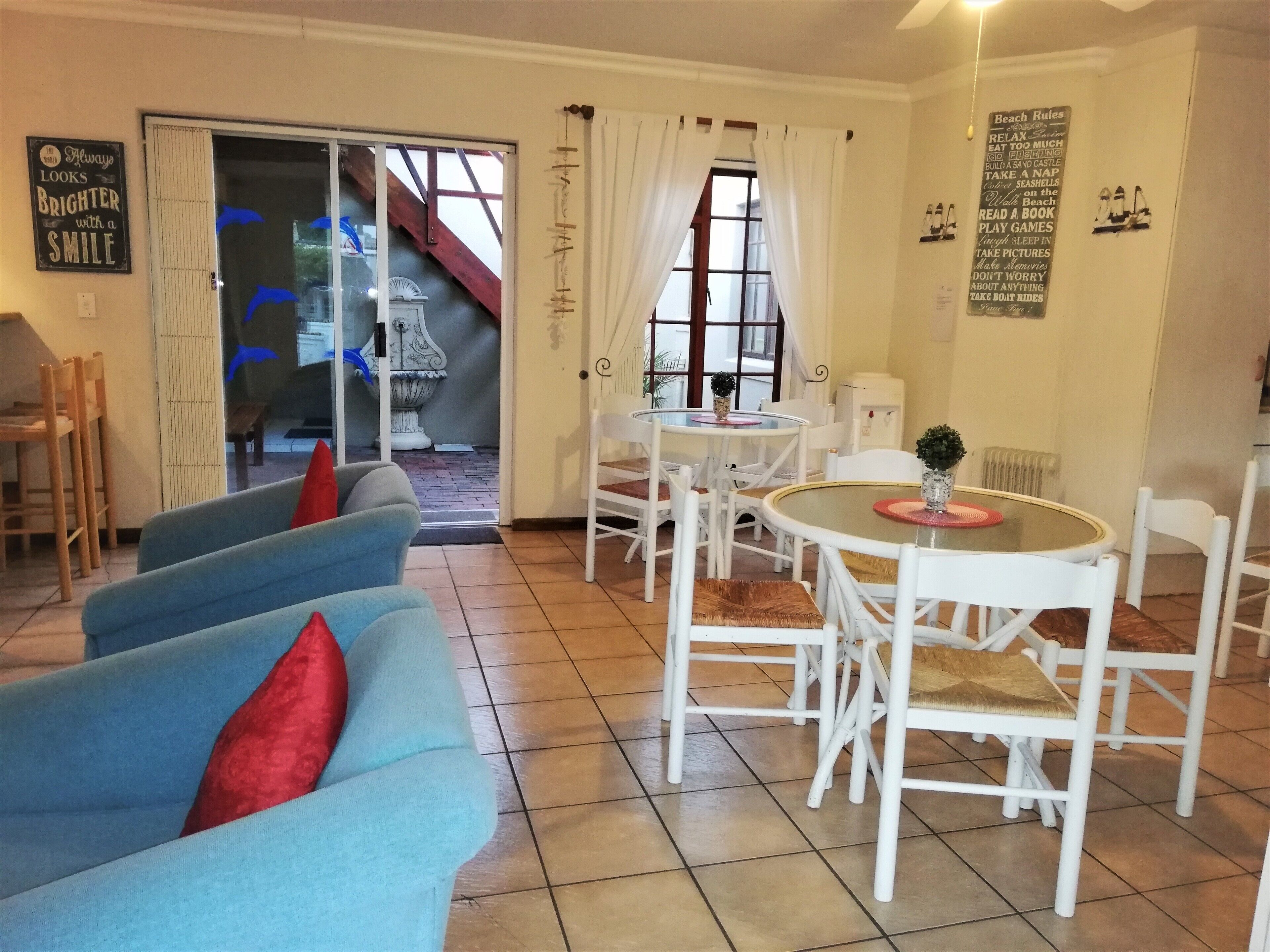 Una sala de estar acogedora con un sofá azul, sillas blancas y una pequeña mesa redonda. Una escalera lleva a un balcón, y un cartel con normas de la playa es visible en la pared.