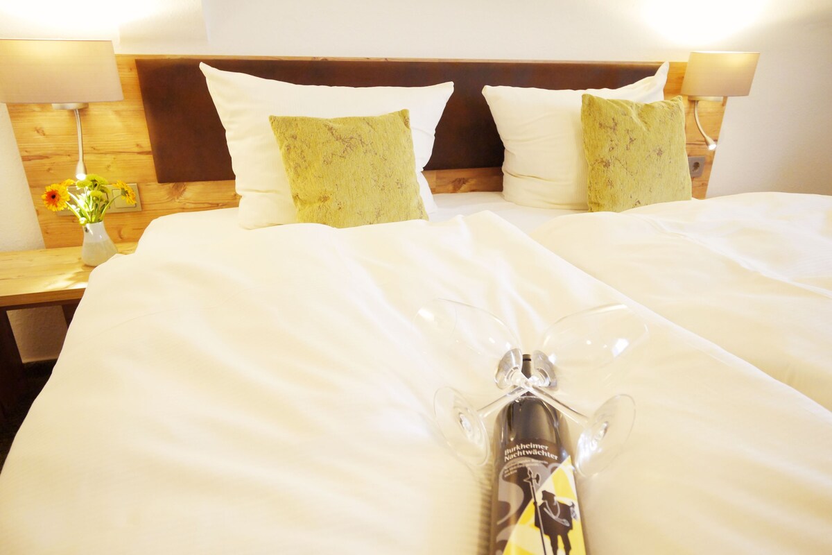 Una habitación de hotel con una cama bien hecha, dos almohadas, una mesita de noche con lámpara y un jarrón con flores.