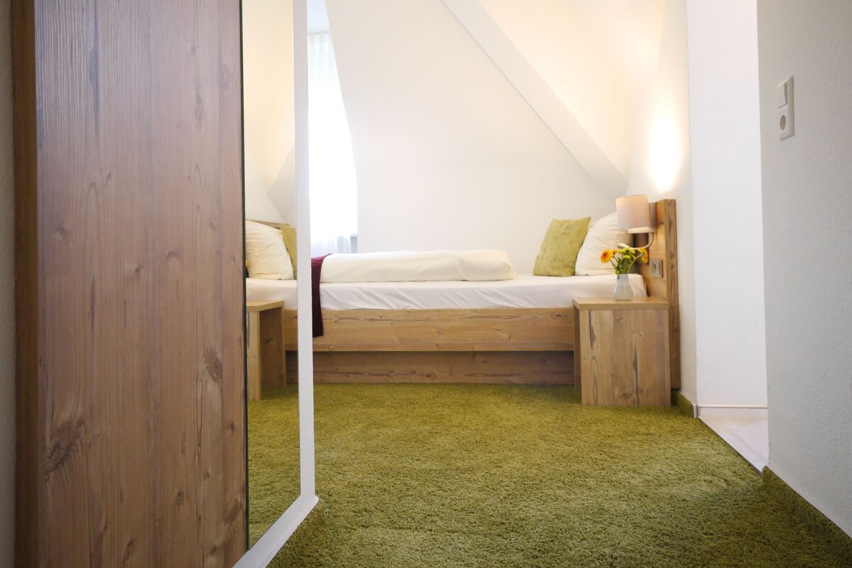 Un dormitorio con techo inclinado, una cama con ropa de cama blanca, una mesita de noche con lámpara y un pequeño florero con flores.