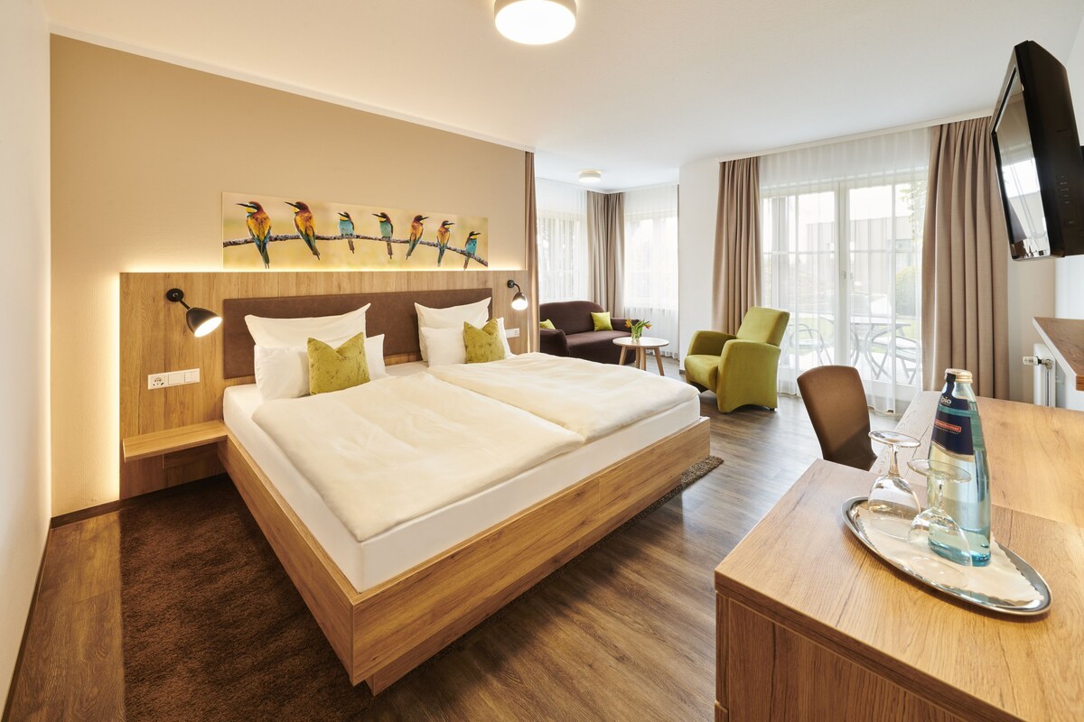 Habitación de hotel con una cama grande, un escritorio con una botella y vasos, un televisor y un cuadro de pájaros en la pared.