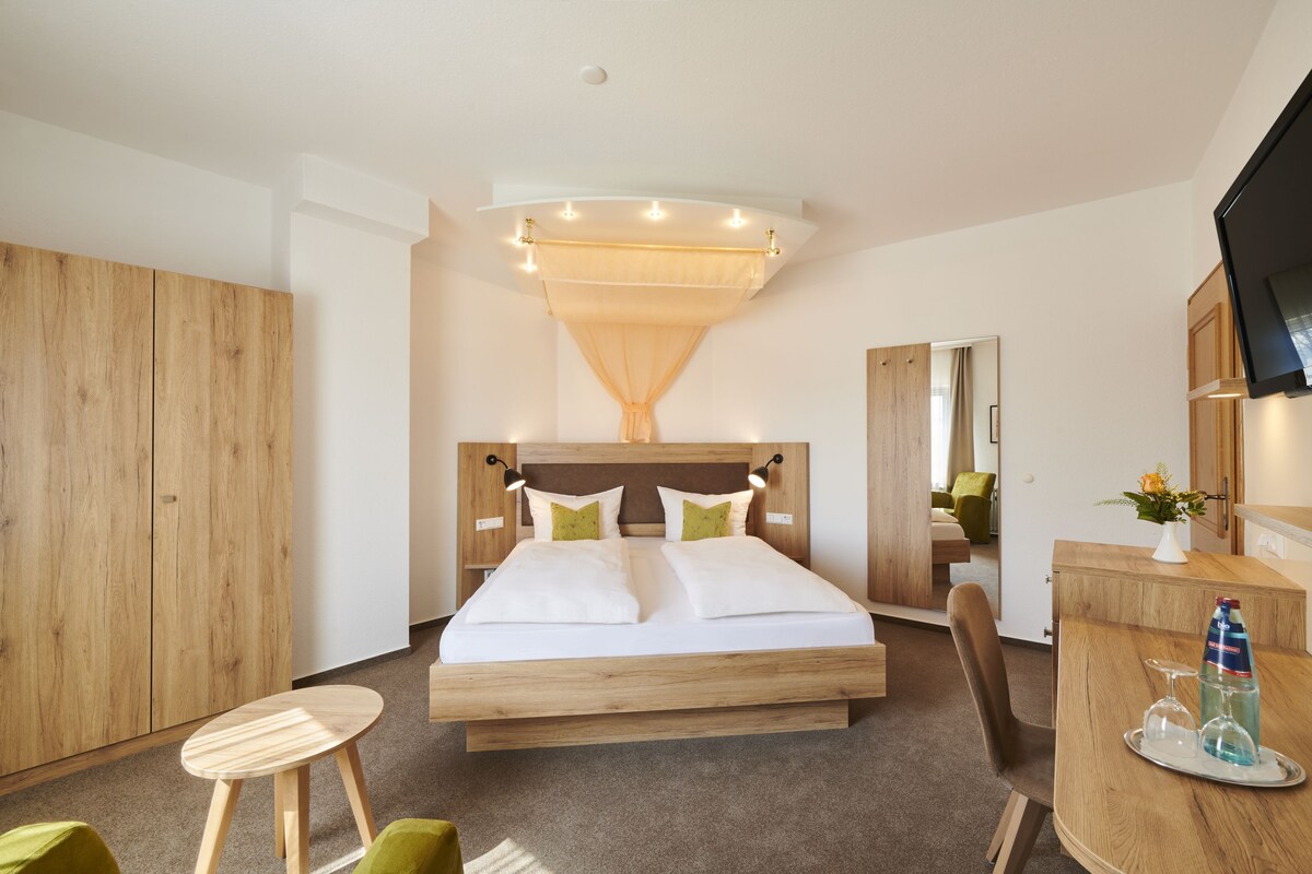 Habitación de hotel moderna con una cama de madera, un escritorio con una botella y vasos, y un televisor en la pared.
