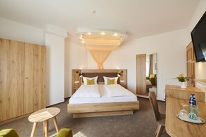 Quarto de hotel moderno com cama de madeira, escrivaninha com garrafa e copos, e televisão na parede.