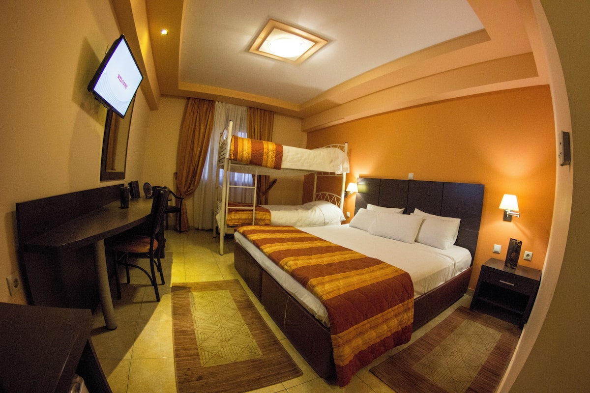 Quarto de hotel com beliche, escrivaninha com cadeira, televisão e mesa de cabeceira com abajur.
