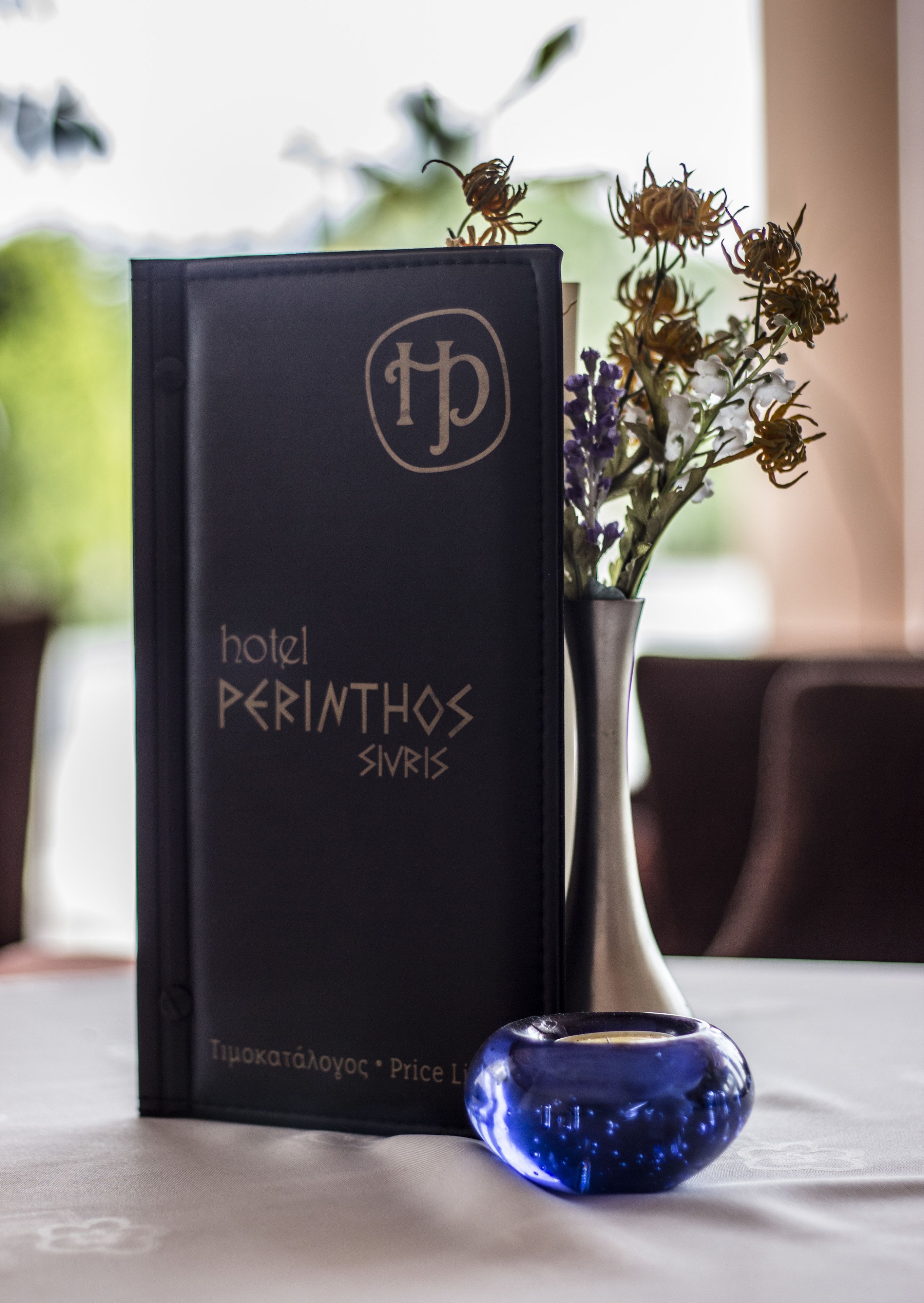 Un folleto de hotel negro con letras doradas, un jarrón de plata con flores secas y un objeto de vidrio azul sobre una mesa.