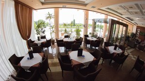 En restaurant med borde og stole arrangeret i en halvcirkel, store vinduer med gardiner og udsigt til grønne områder.