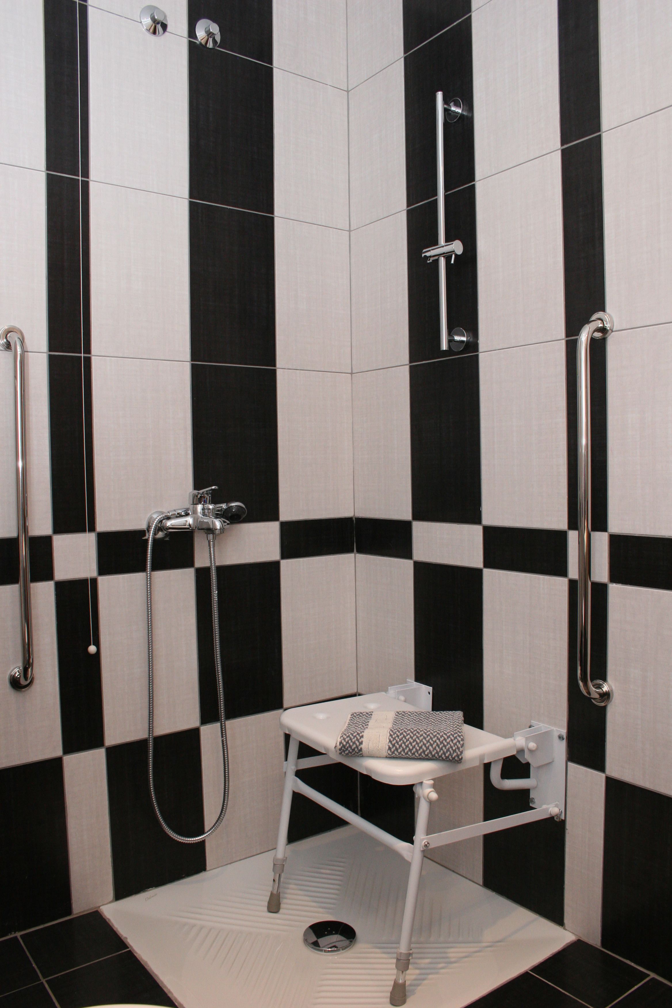 A bathroom with black and white checkered tiles, a shower with a handheld showerhead, and a white bench with a towel.