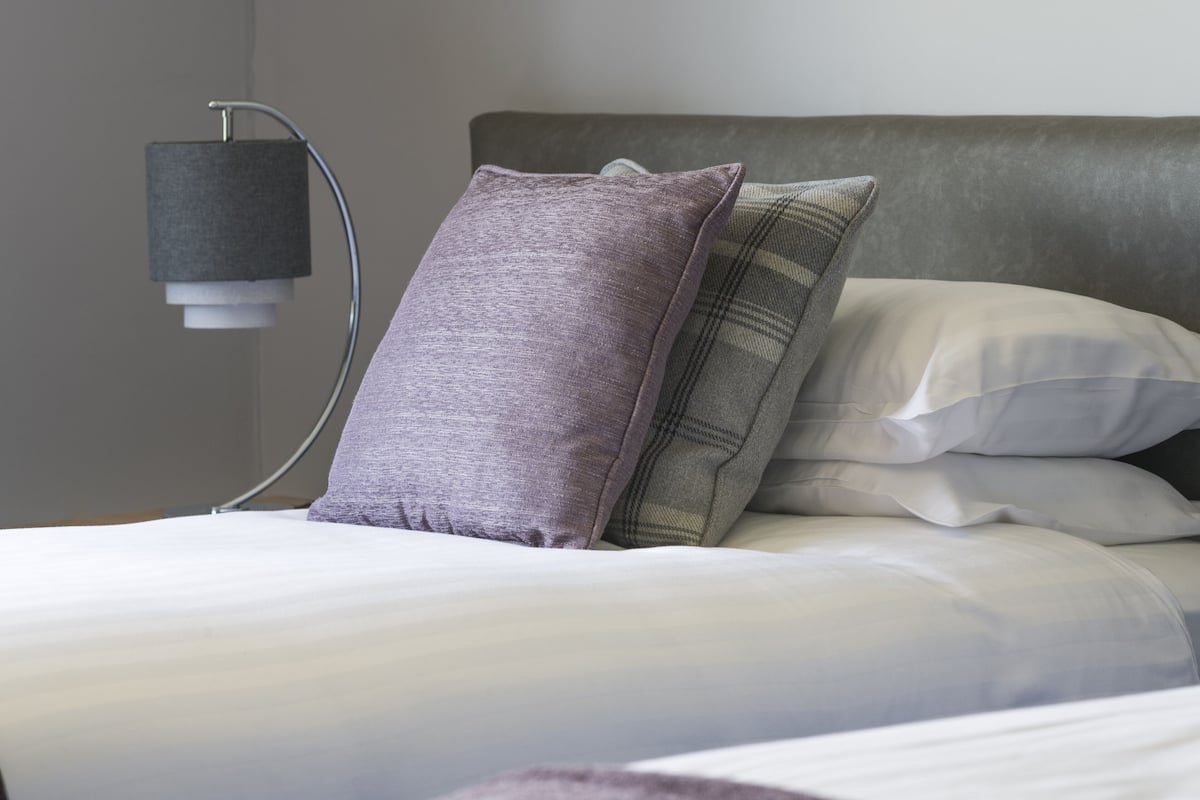 A neatly made bed with a grey headboard, white bedding, and a purple pillow.
