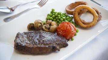 Une assiette contenant un steak, des champignons, des petits pois, une tomate et des rondelles d’oignon.