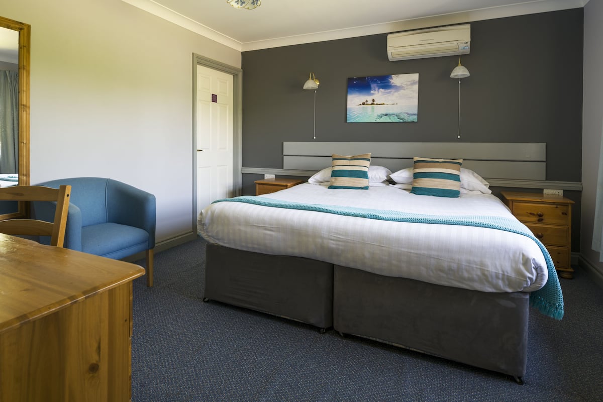 A bedroom with a large bed, a wooden desk, a blue chair, and a nightstand with a lamp.