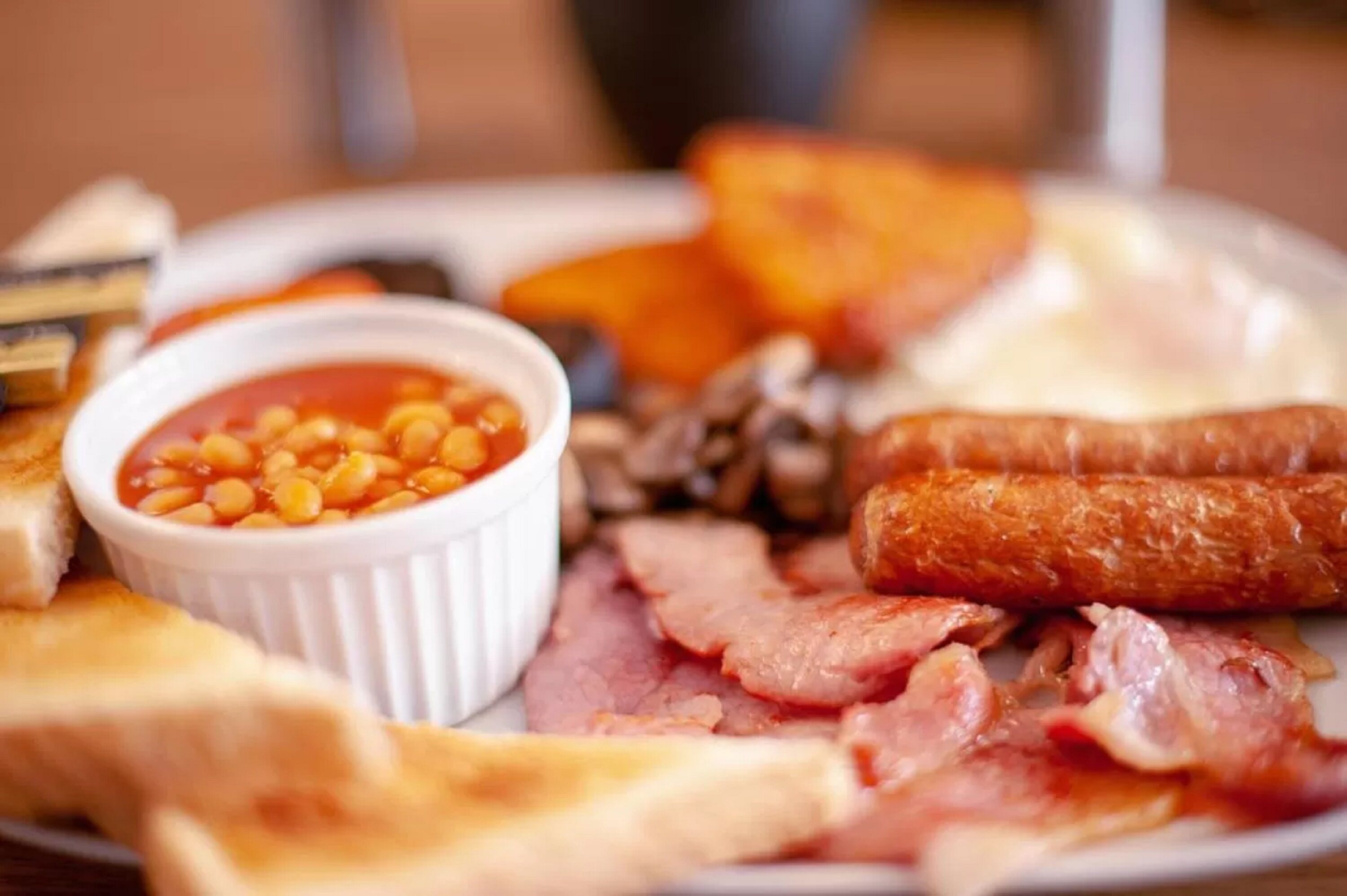 A breakfast plate with toast, beans, bacon, sausages, and a small bowl of sauce.