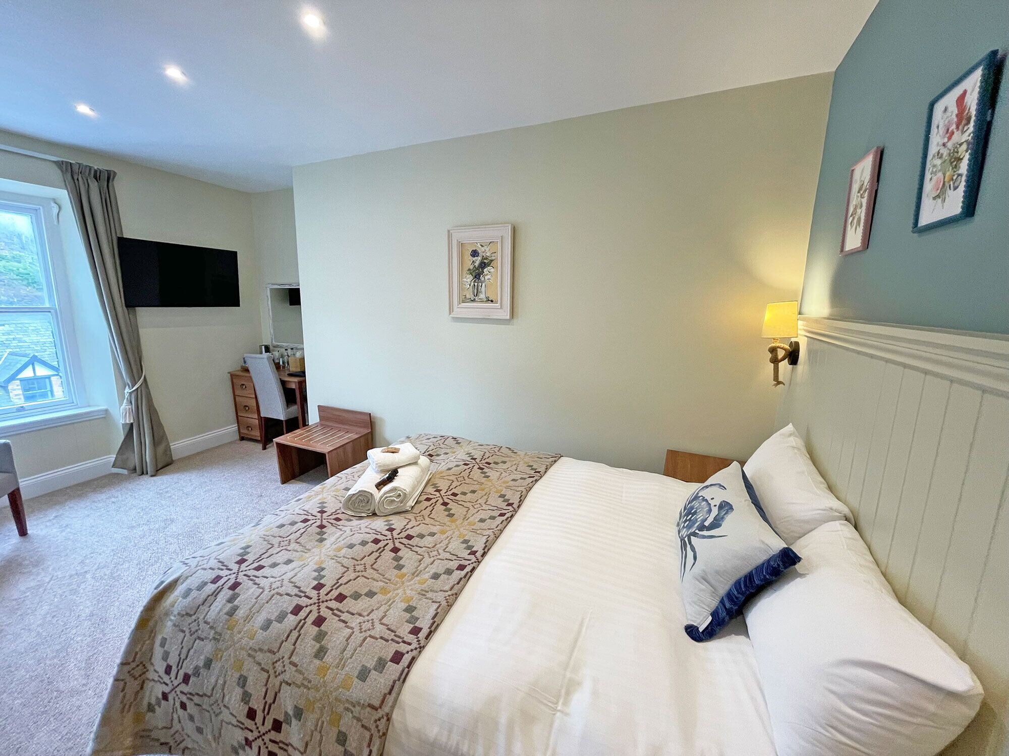 A bedroom with a bed, a desk, a chair, a TV, and a window with curtains.