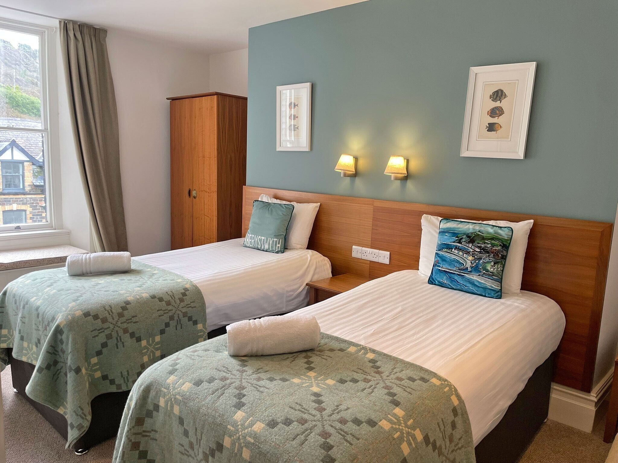 A hotel room with two beds, a wardrobe, a window with curtains, wall-mounted lights, and framed pictures.