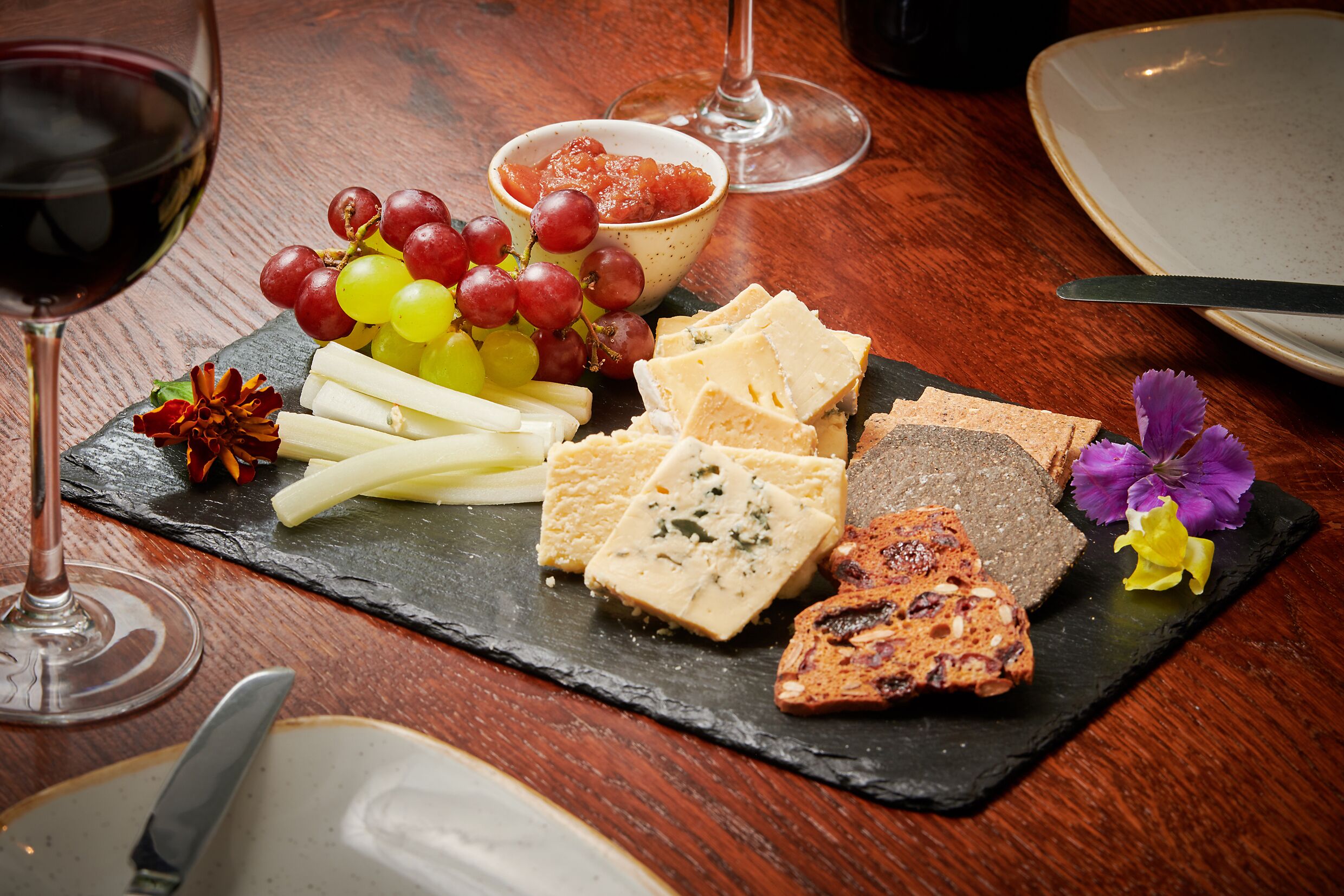 A slate board with various cheeses, grapes, and a bowl of fruit preserves.