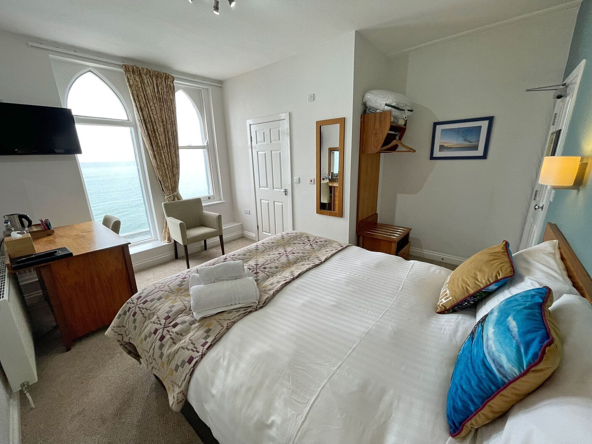 A hotel room with a bed, a desk, a chair, a window with curtains, a mirror, and a picture on the wall.