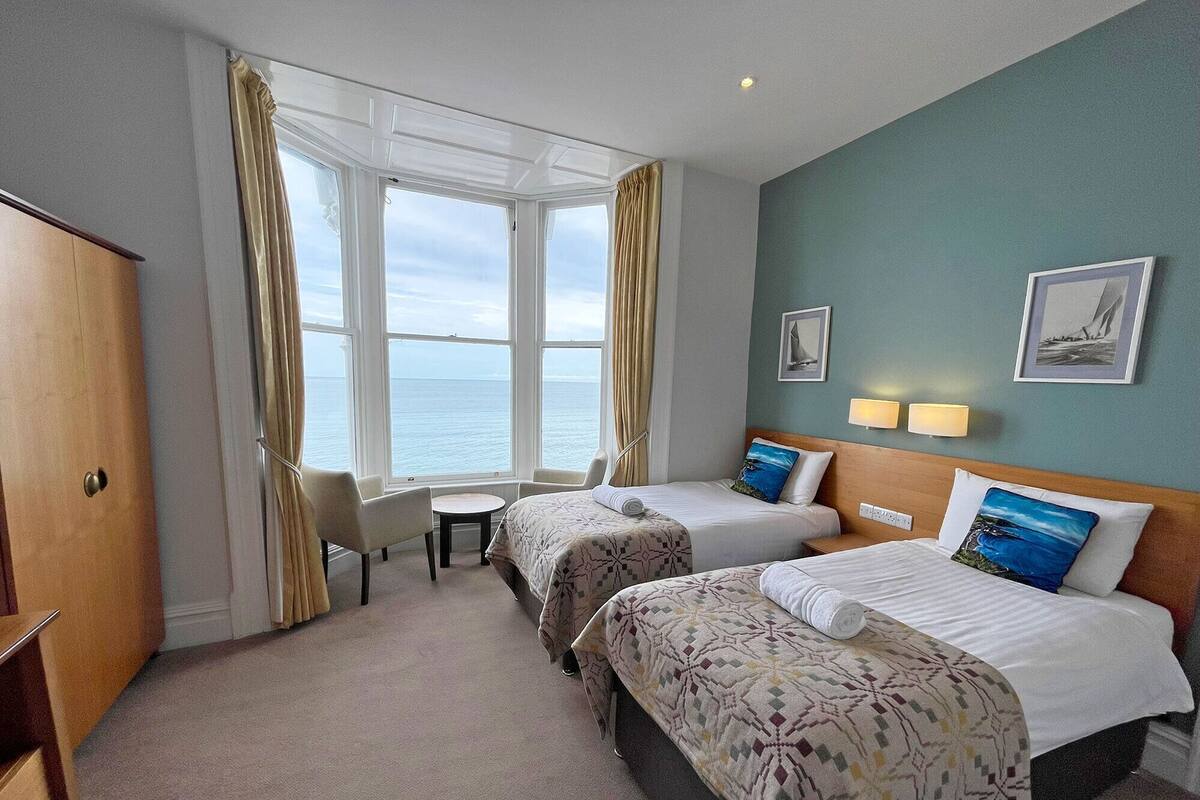 A hotel room with two beds, a desk, and a view of the sea.