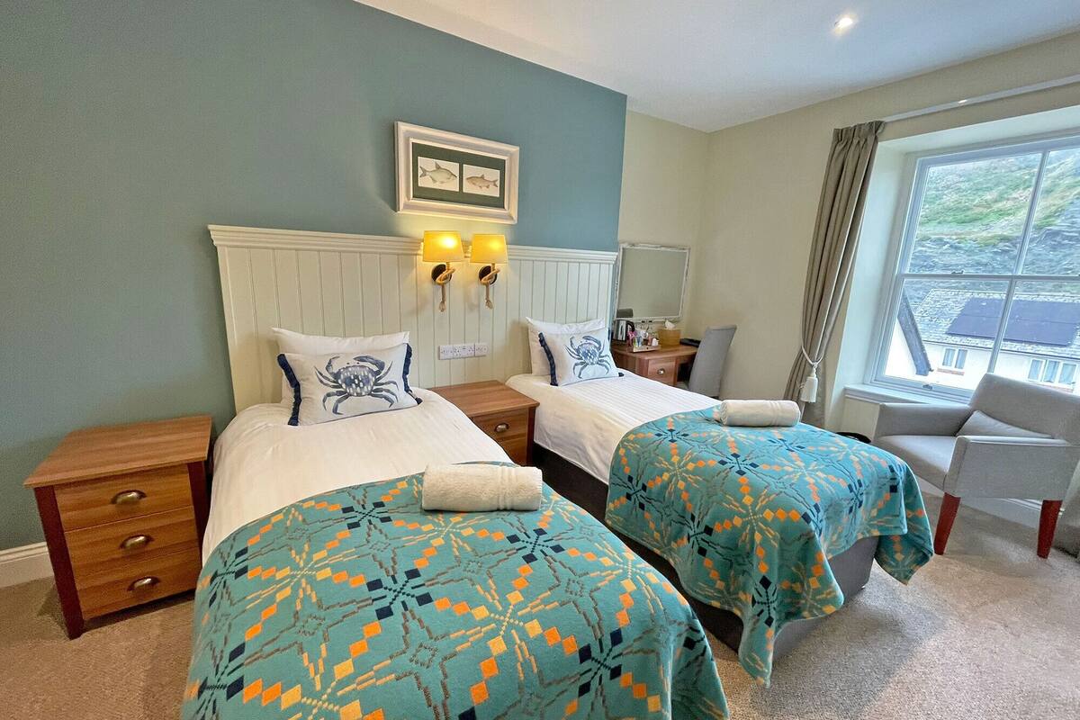 A hotel room with two beds, a window with a view, and a small desk with a lamp.