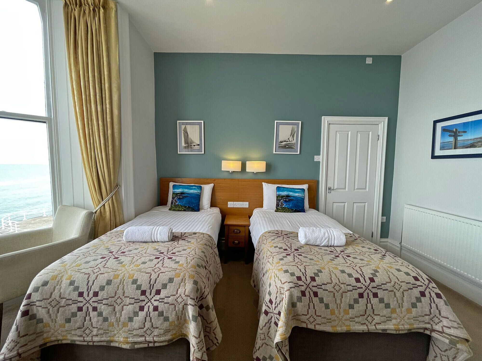 A hotel room with two beds, a window with curtains, a door, and wall-mounted lamps.