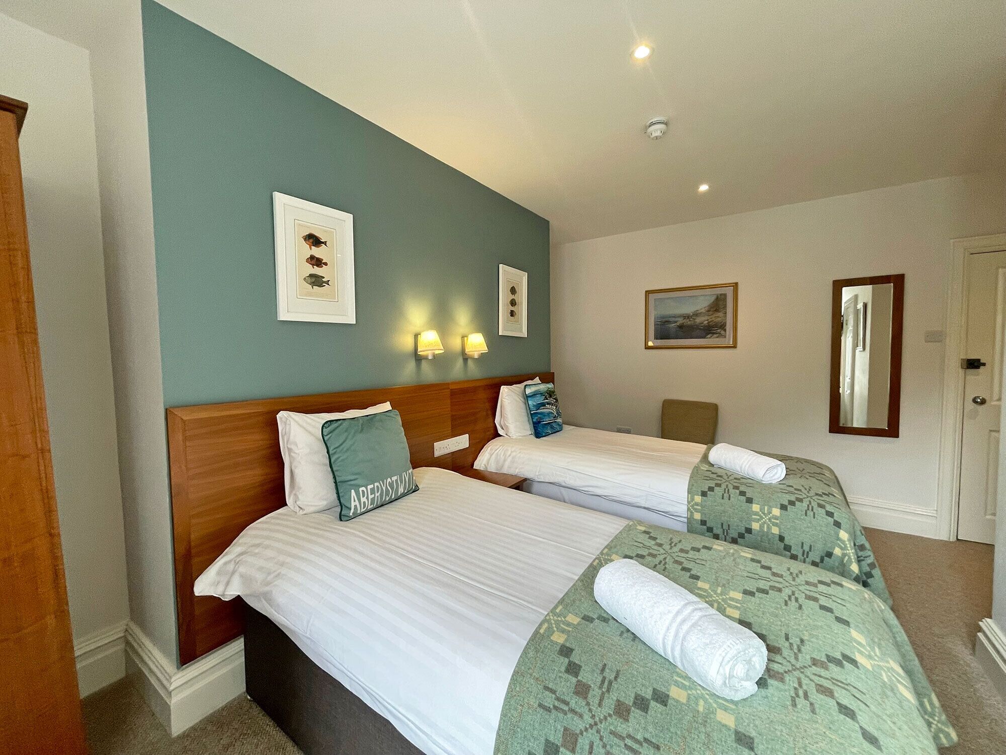 A hotel room with two beds, a wooden headboard, a teal accent wall, and wall-mounted lights.