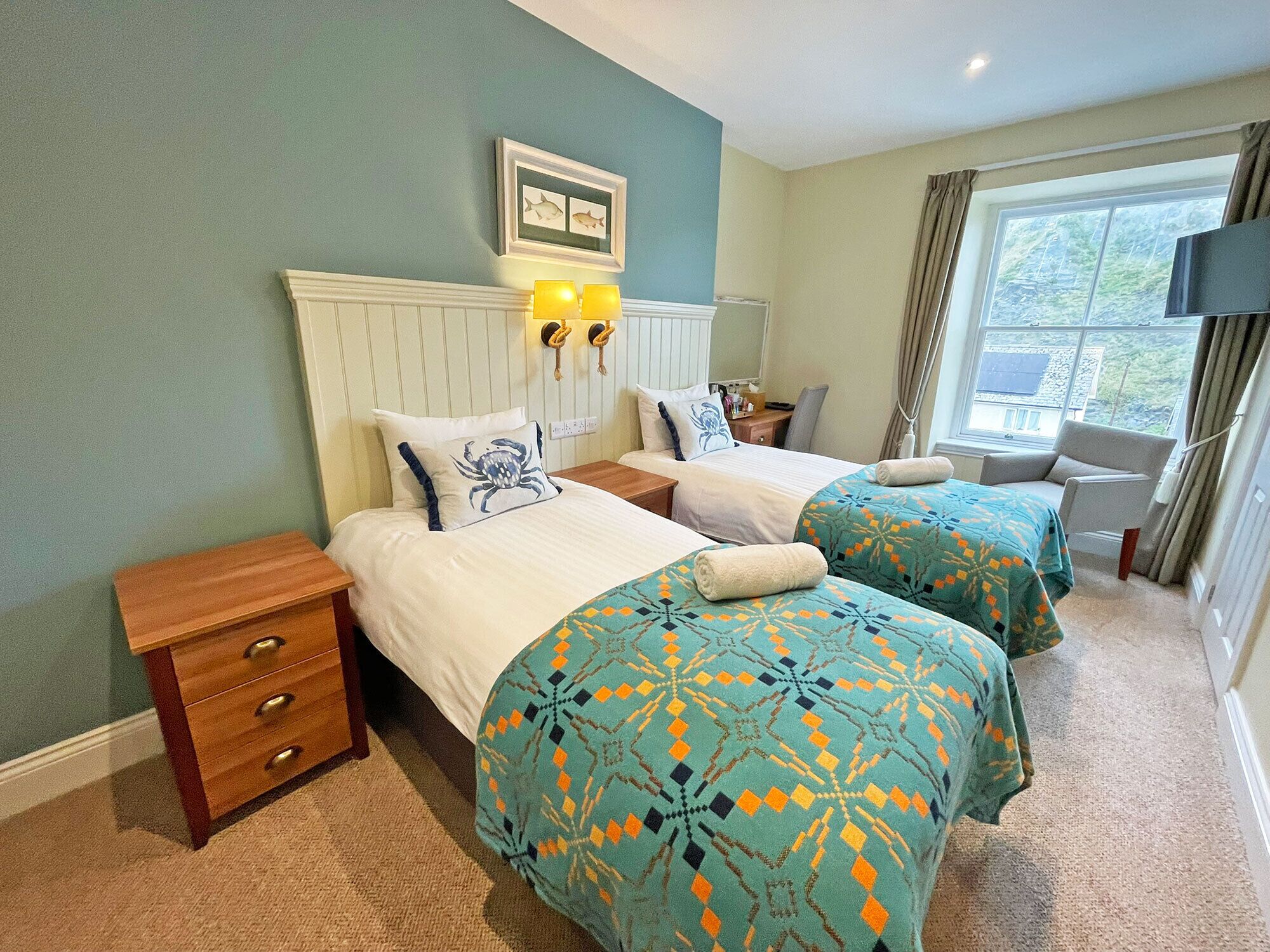A hotel room with two beds, a wooden nightstand, a dresser, a window with curtains, and a television.