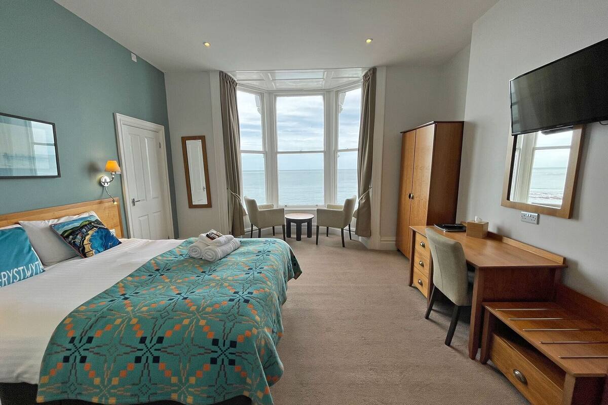 A hotel room with a bed, a desk, a chair, a TV, and a view of the sea.