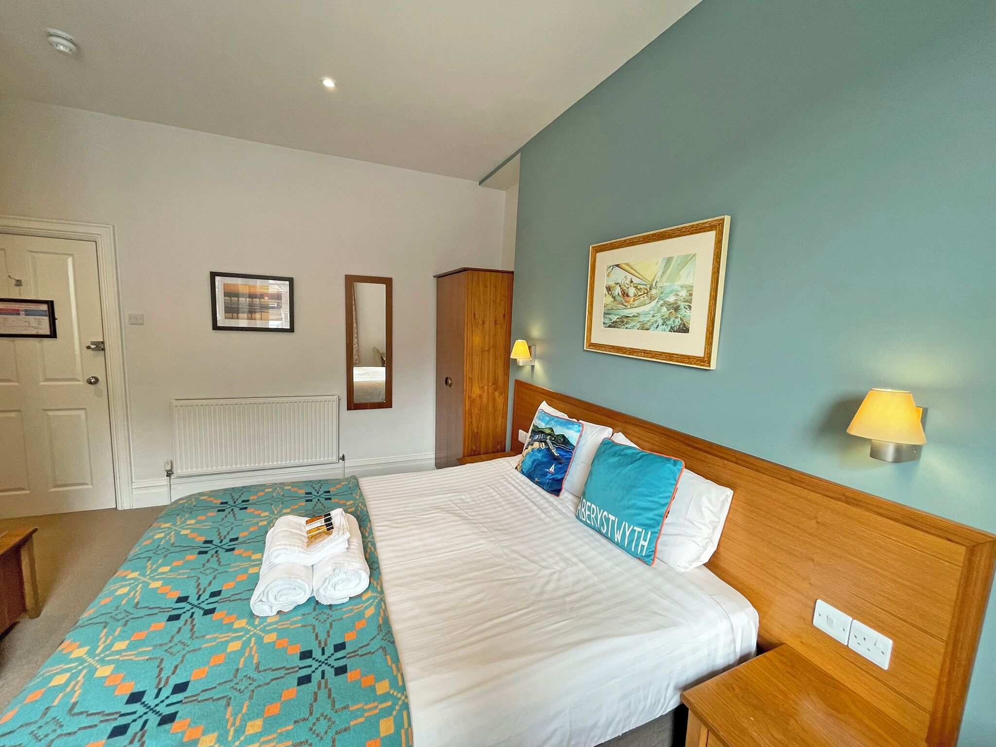 A hotel room with a bed, a wooden wardrobe, a radiator, a framed picture, and a wall-mounted lamp.