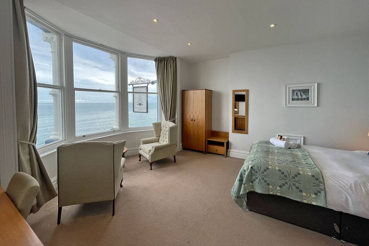 A hotel room with a large window offering a sea view, a bed with a patterned bedspread, a chair, a wardrobe, and a small table.