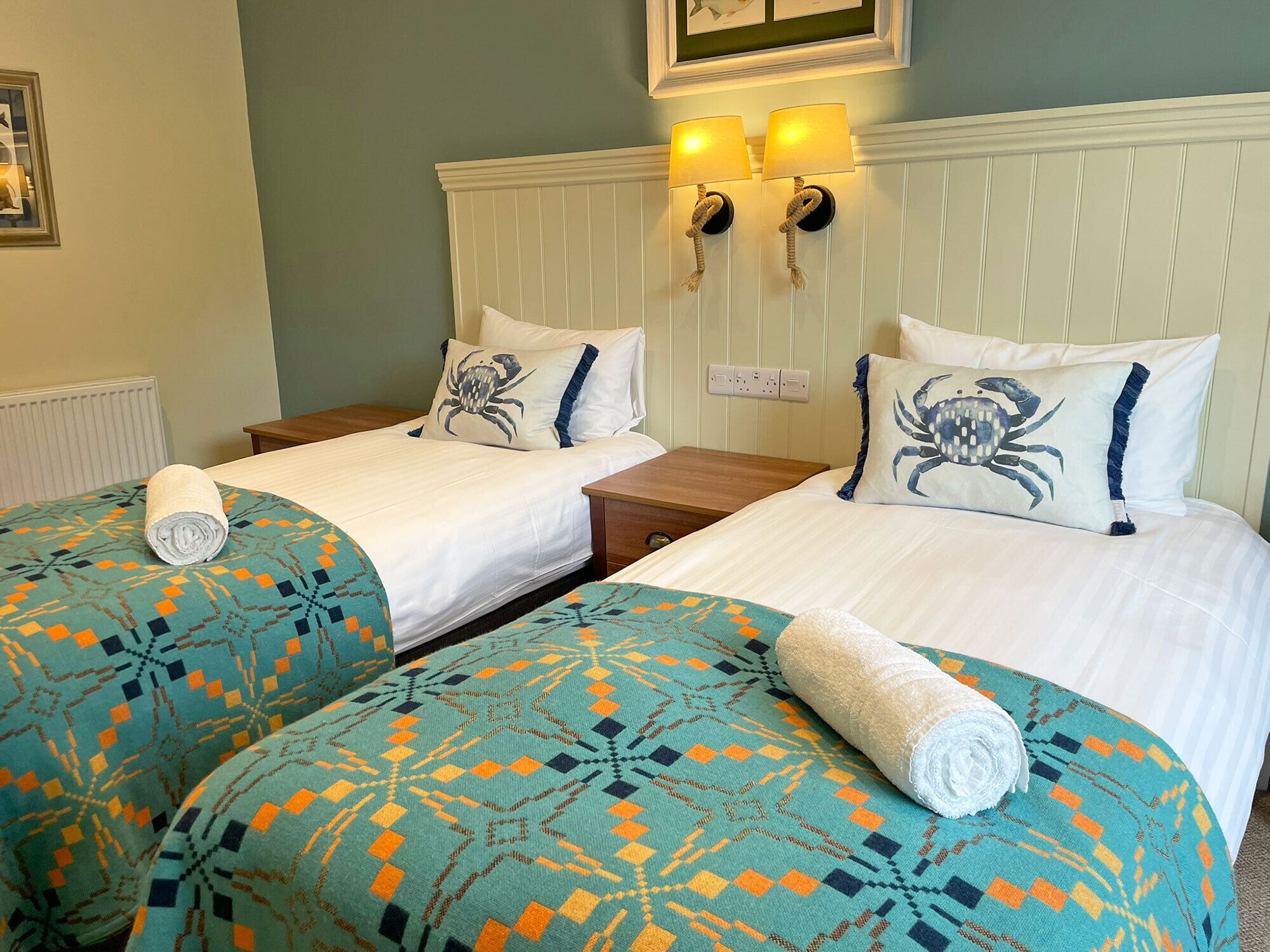 A hotel room with two beds, each with a patterned bedspread and matching pillows, a white headboard, and a wall-mounted lamp.