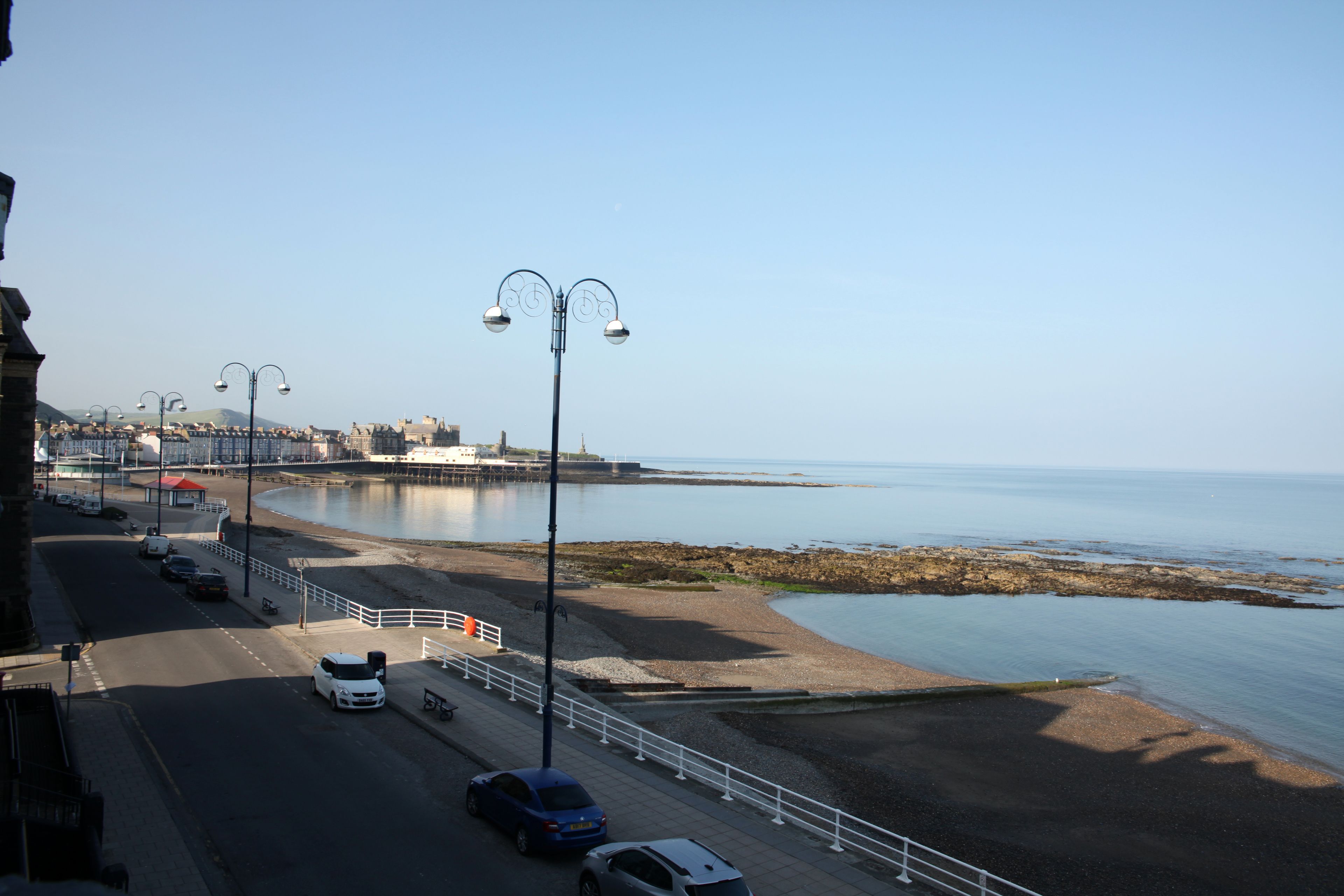 A coastal road with parked cars, a pedestrian walkway, and a clear view of the sea.