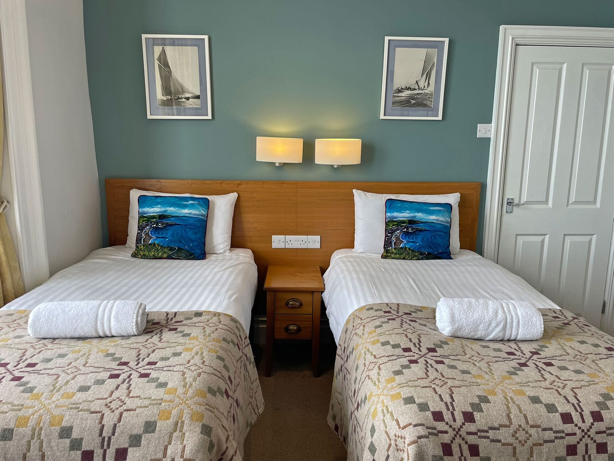 A hotel room with two beds, a wooden headboard, a nightstand, a lamp, and two framed pictures on the wall.