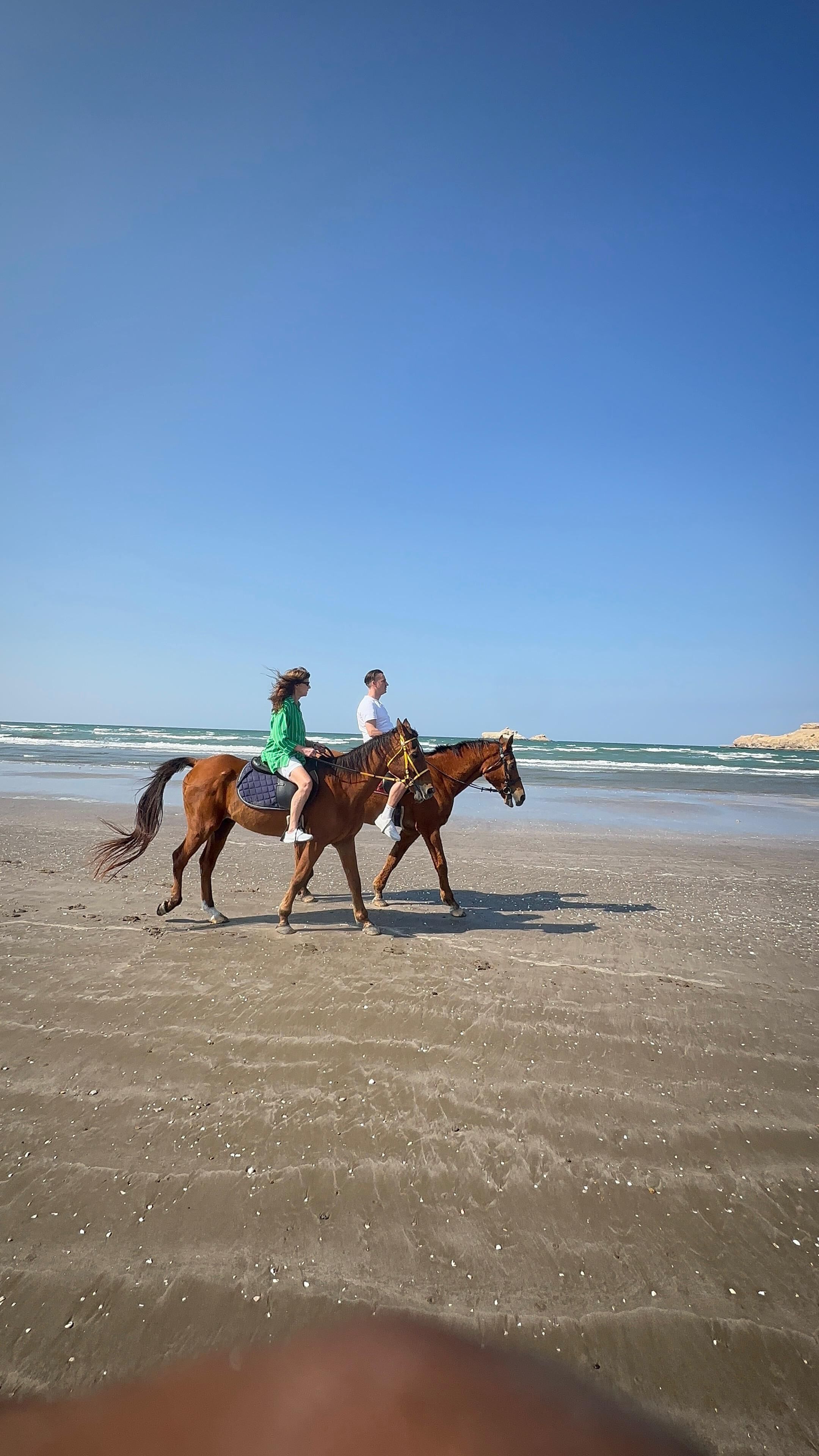Deux personnes à cheval sur une plage, avec l’océan en arrière-plan.