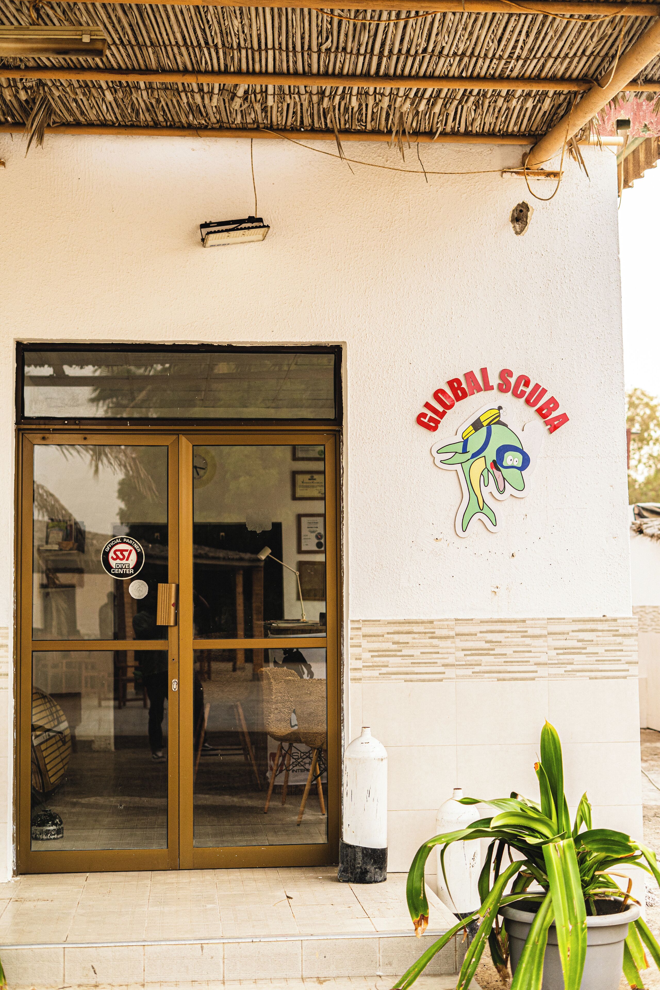 Entrada de un edificio con puerta de vidrio, un cartel que dice GLOBAL SCUBA y una planta en maceta al frente.