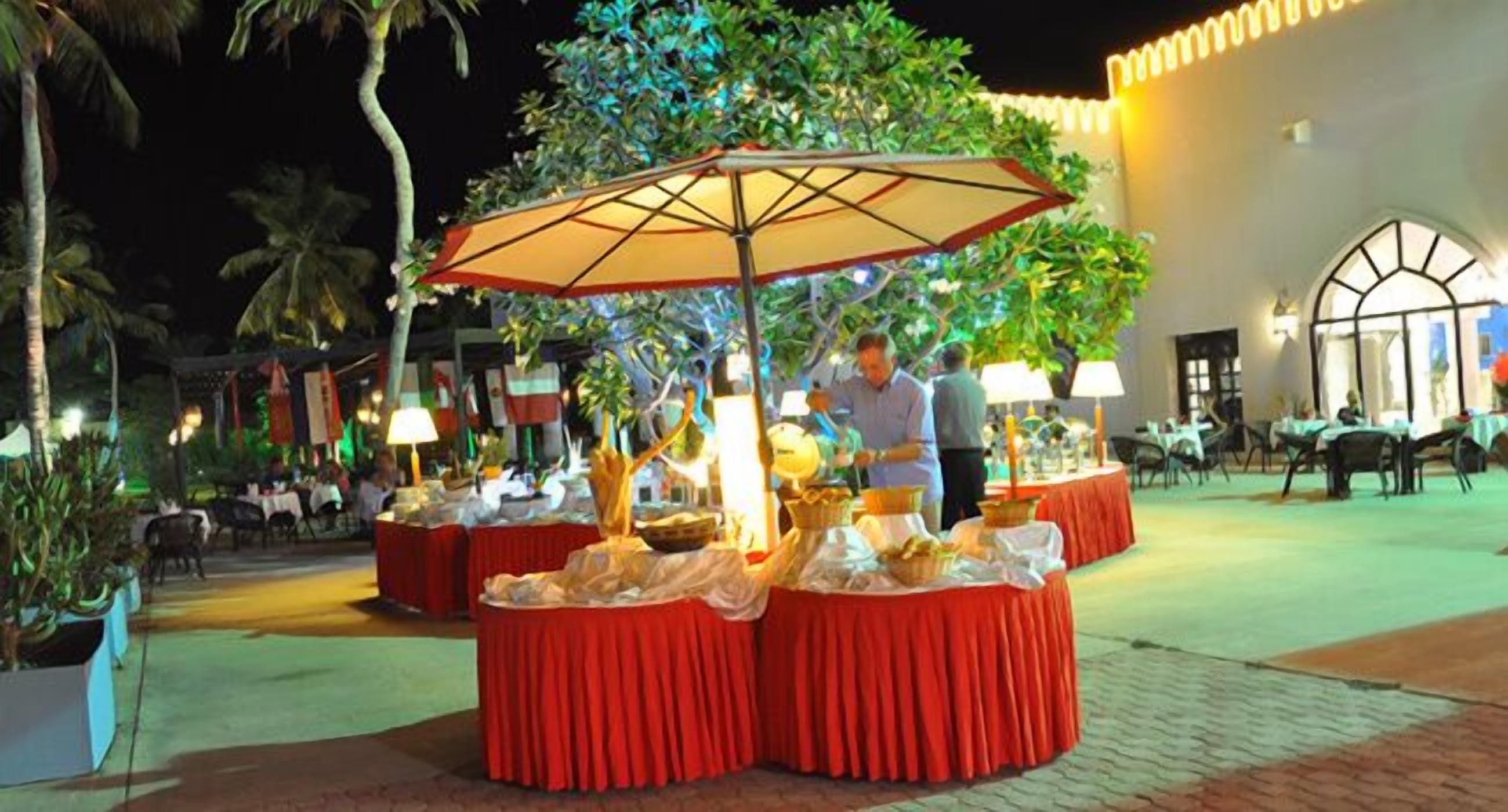 An outdoor dining area with round tables covered in red tablecloths, a central umbrella, and a person standing nearby.