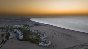 Vista aérea de un complejo turístico costero con villas frente a la playa, una playa de arena y un paisaje urbano al fondo.