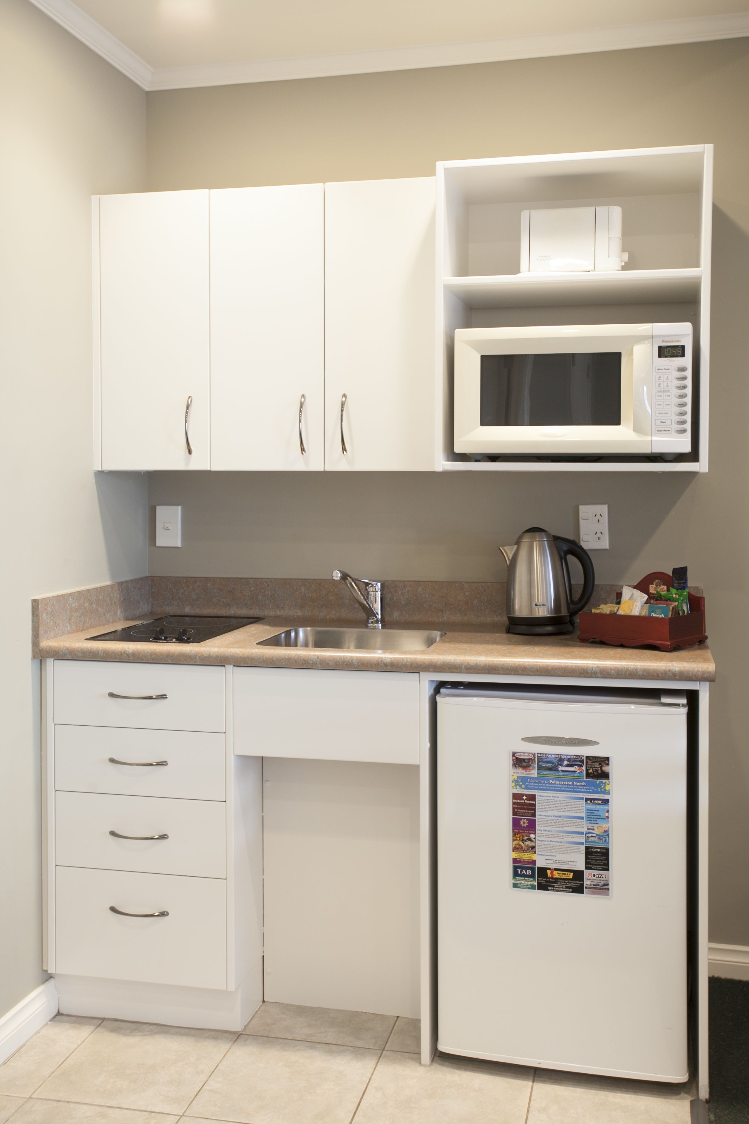 A compact kitchen with white cabinets, a microwave, a kettle, and a refrigerator.