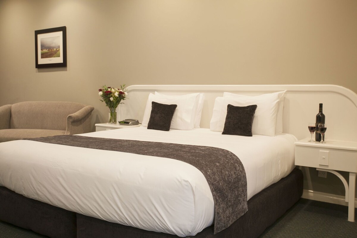 A hotel room with a large bed, a chair, a nightstand with a bottle and glass, and a framed picture on the wall.