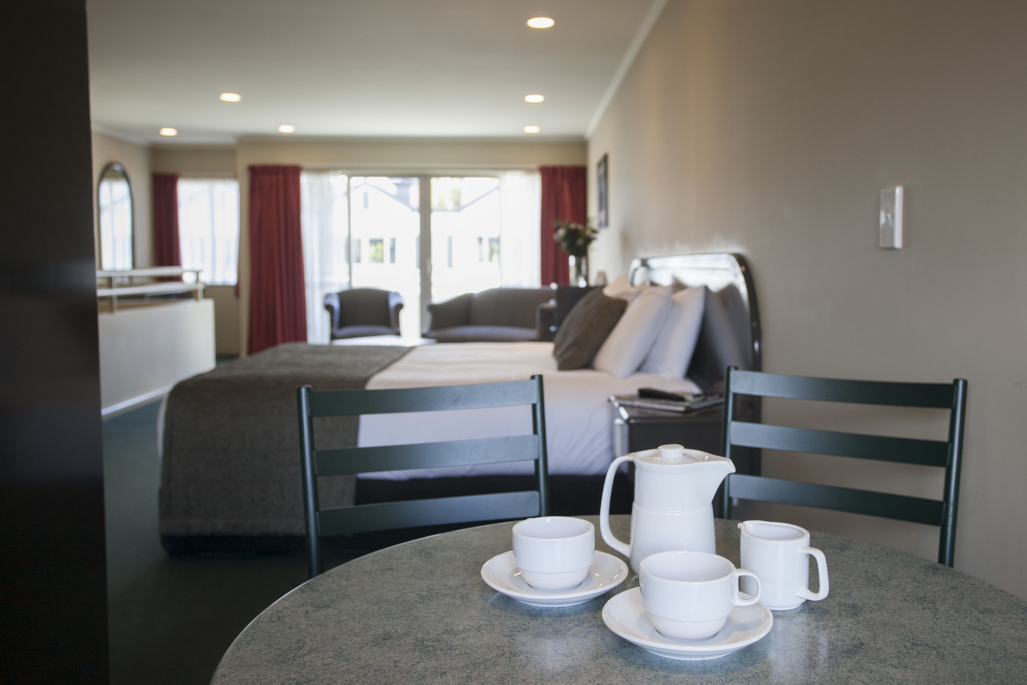 A hotel room with a bed, two chairs, a table with cups and a pitcher, and a view of the outside.