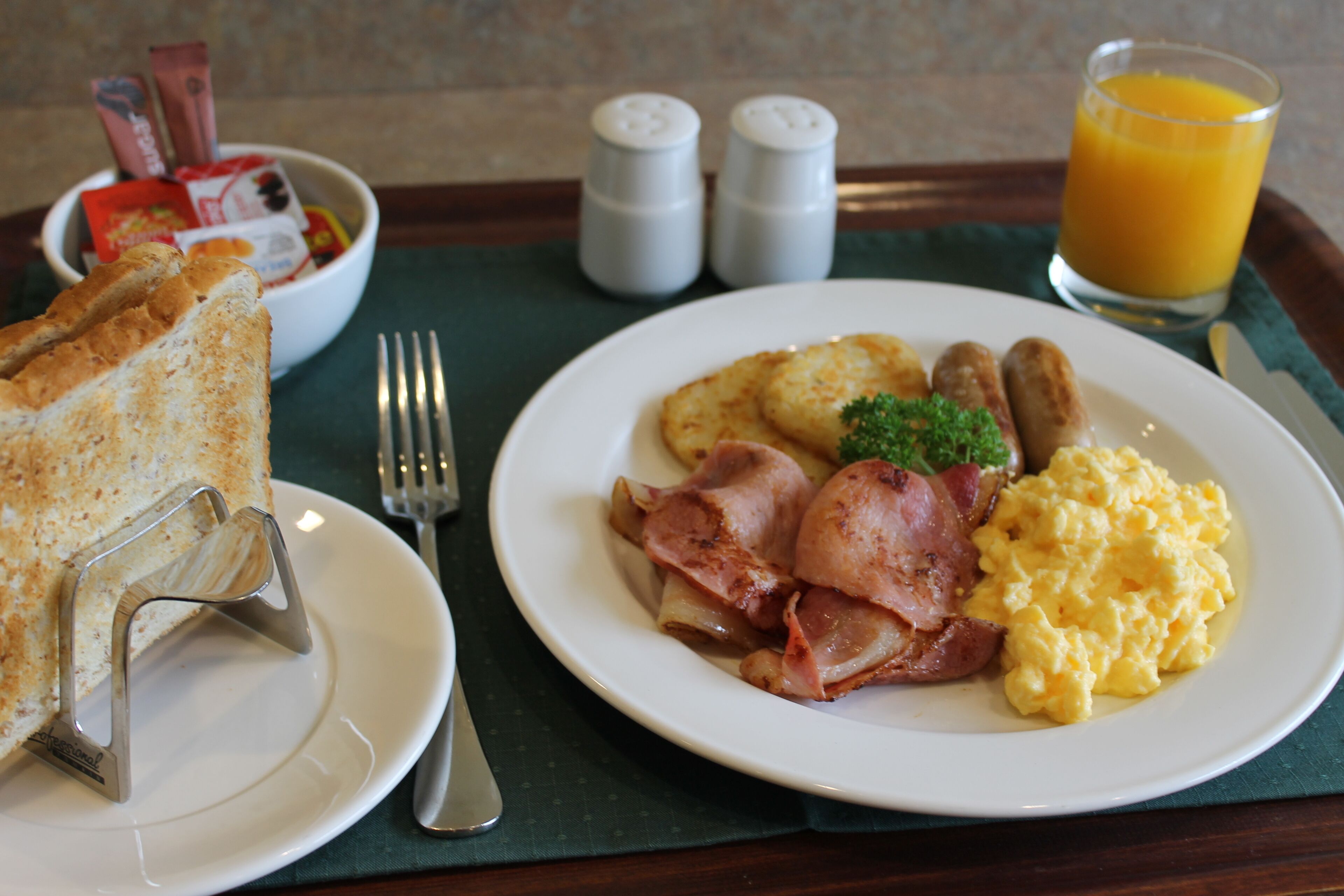 A breakfast tray with toast, scrambled eggs, sausages, and ham, along with a glass of orange juice and a bowl of cereal.