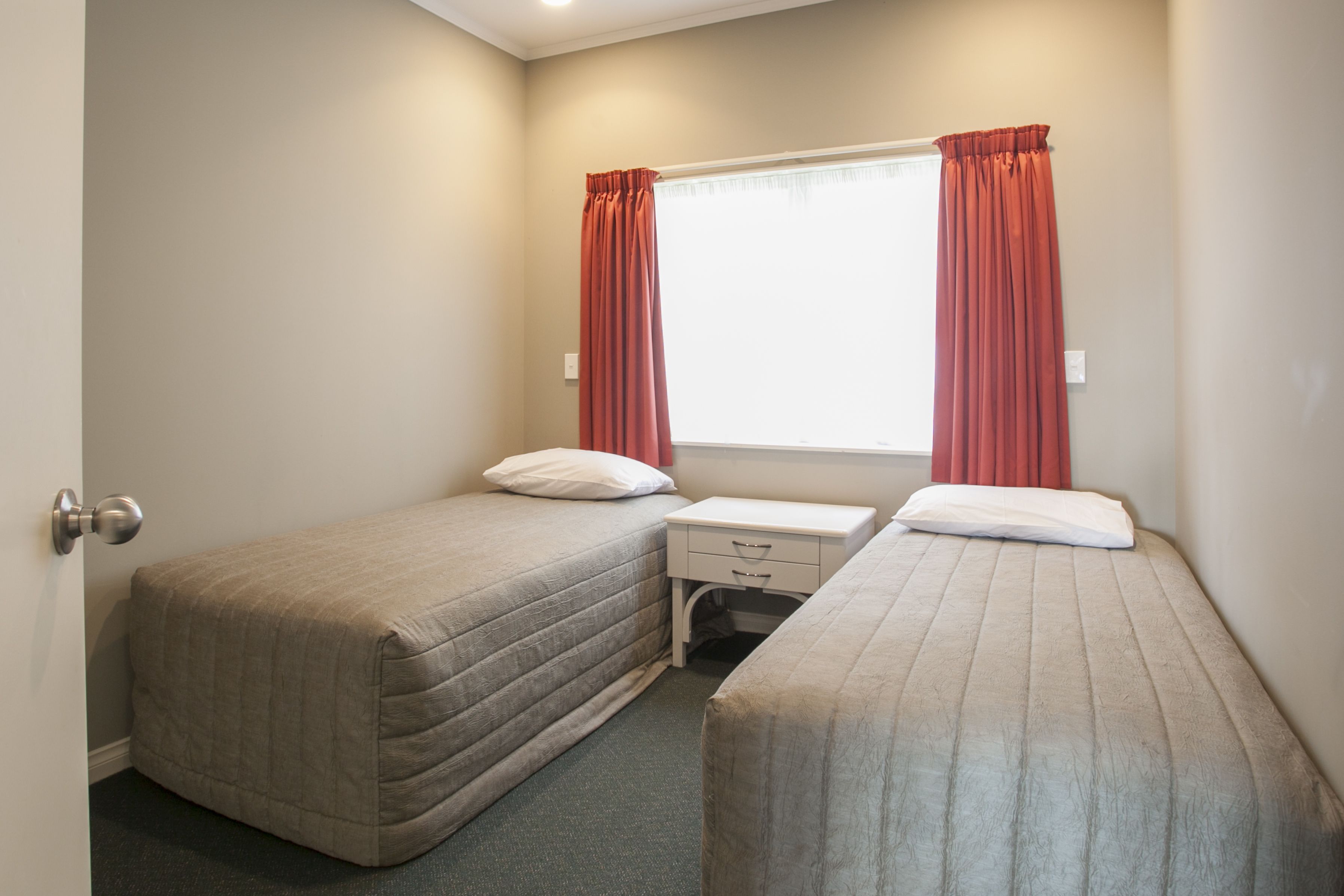 A small, clean room with two single beds, a window with curtains, a bedside table, and a door.