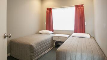 A small, clean room with two single beds, a window with curtains, a bedside table, and a door.