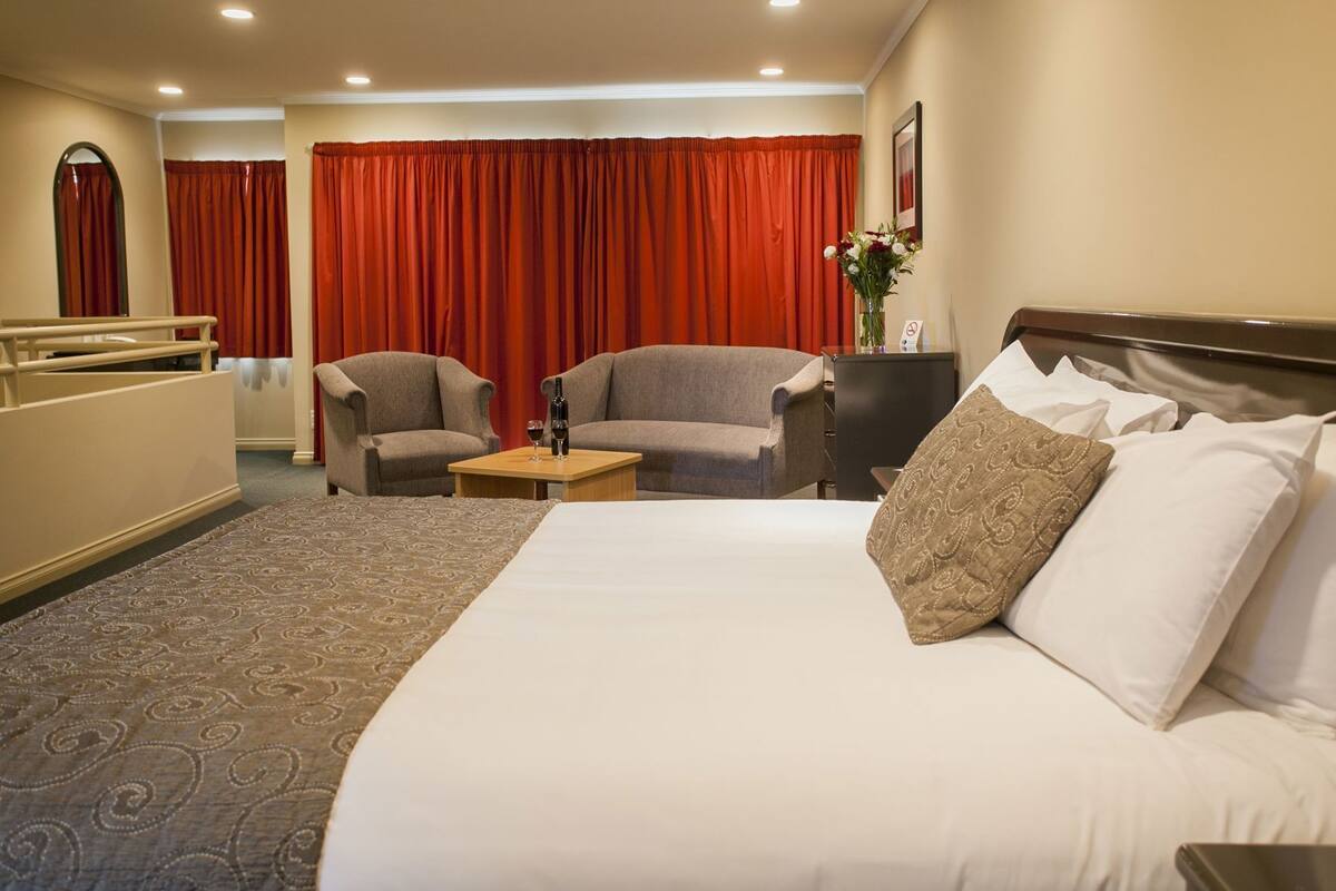Ein Hotelzimmer mit einem großen Bett, zwei Sesseln, einem kleinen Tisch mit einer Flasche und einem Glas sowie einem roten Vorhang im Hintergrund.