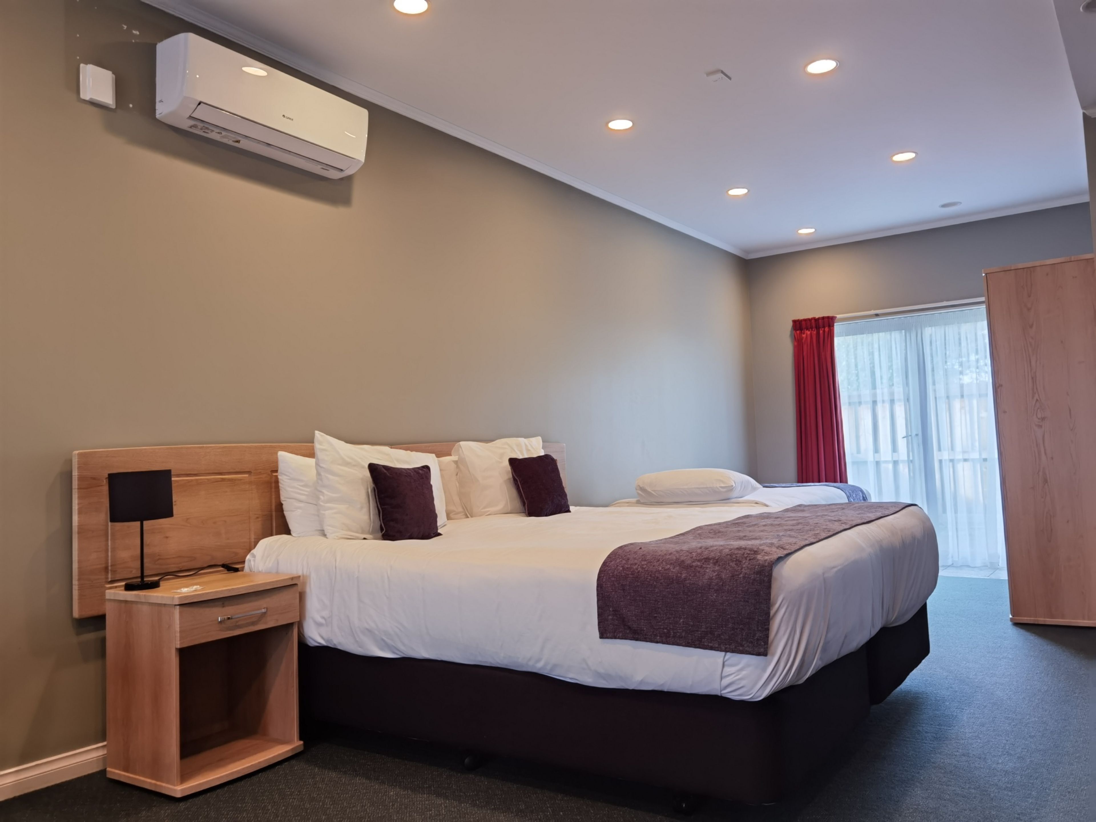 A modern hotel room with a large bed, a bedside table, a lamp, an air conditioning unit, and a window with curtains.