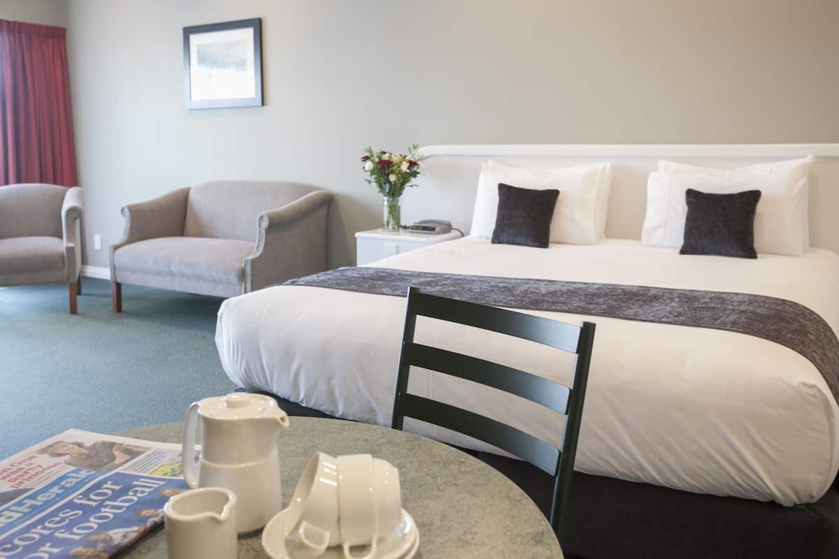 Ein Hotelzimmer mit einem Bett, einem Sessel, einem Tisch mit einer Zeitung, einer Vase mit Blumen und einem gerahmten Bild an der Wand.
