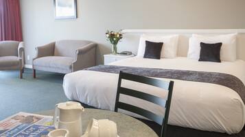 Un cuarto de hotel con una cama, una silla, una mesa con un periódico, un florero con flores y un cuadro en la pared.